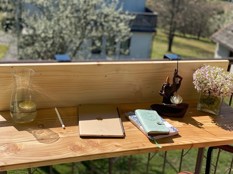 May include: A wooden desk with a laptop, two notebooks, a glass of water, a wooden incense holder, and a vase of pink flowers. The desk is on a balcony with a view of trees and a building in the background.