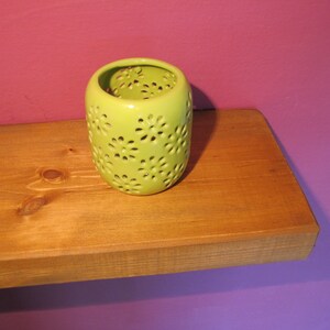 Może przedstawiać: Limonkowo-zielony ceramiczny świecznik z wycięciami w kształcie stokrotek. Świecznik stoi na drewnianej p&oacute;łce na tle fioletowej ściany. Świecznik jest cylindryczny i ma błyszczące wykończenie.