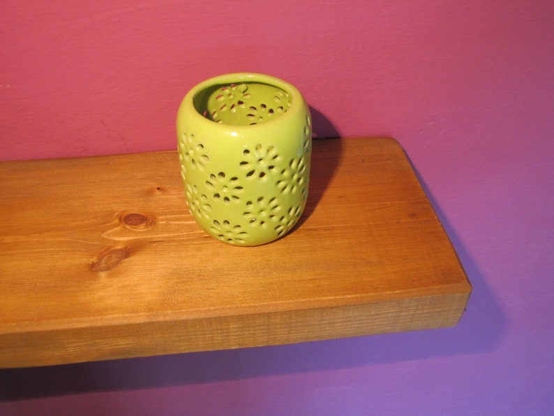 Może przedstawiać: Limonkowo-zielony ceramiczny świecznik z wycięciami w kształcie stokrotek. Świecznik stoi na drewnianej p&oacute;łce na tle fioletowej ściany. Świecznik jest cylindryczny i ma błyszczące wykończenie.