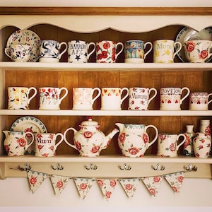 Peut inclure: Une étagère en bois blanc à deux niveaux, décorée d'une variété de tasses, de théières et d'autres articles de cuisine. L'étagère a un bord festonné et est décorée d'un motif floral. Les tasses sont de couleurs et de motifs différents, et certaines portent du texte, comme "Mum" et "Love Joy".