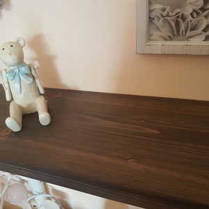 Puede incluir: Una estanter&iacute;a de madera marr&oacute;n oscuro con una figura de oso de peluche de color claro con un lazo azul. Encima, un marco con una impresi&oacute;n floral en blanco y negro. Se ven pompones decorativos y luces de cadena.