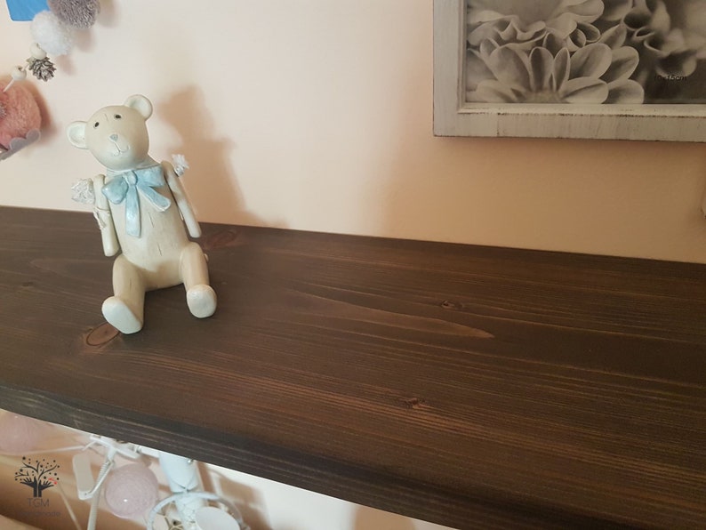 Puede incluir: Una estanter&iacute;a de madera marr&oacute;n oscuro con una figura de oso de peluche de color claro con un lazo azul. Encima, un marco con una impresi&oacute;n floral en blanco y negro. Se ven pompones decorativos y luces de cadena.