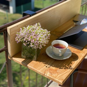May include: A wooden balcony table with a laptop, a cup of tea, and a vase of pink flowers. The table is attached to a railing with metal brackets.