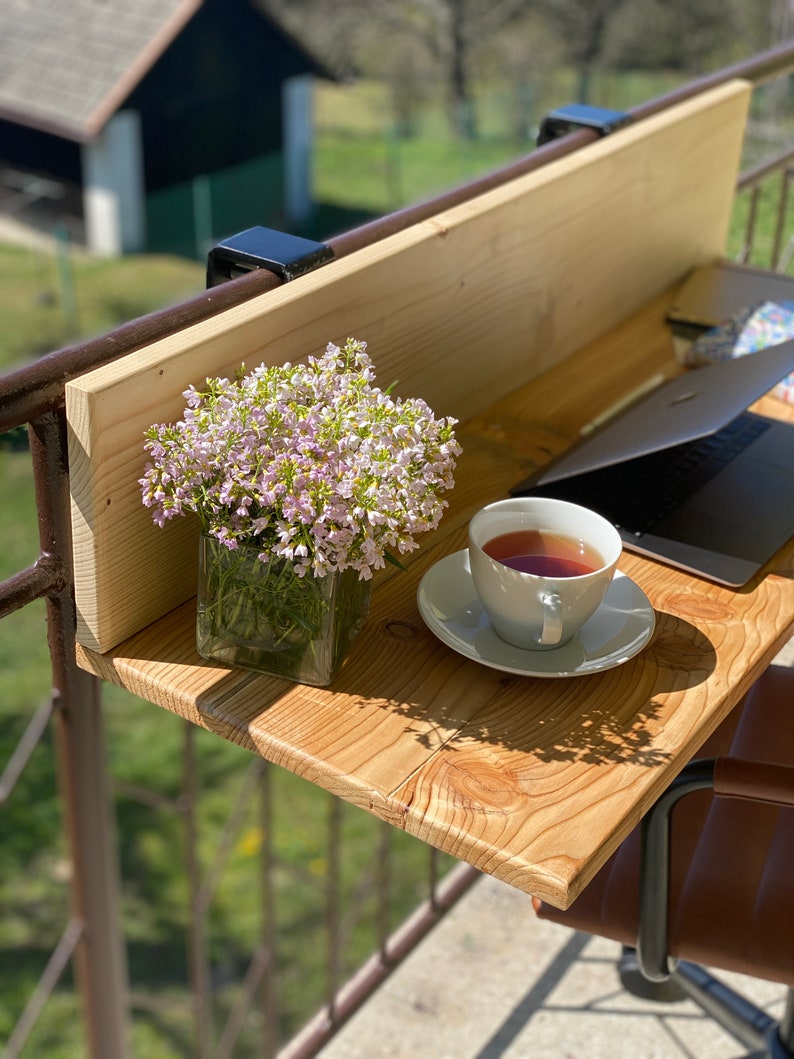 May include: A wooden balcony table with a laptop, a cup of tea, and a vase of pink flowers. The table is attached to a railing with metal brackets.