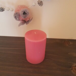 Puede incluir: Una vela rosa en forma de pilar con detalles acanalados verticales. La vela es cil&iacute;ndrica y se encuentra sobre una superficie de madera oscura. Una guirnalda decorativa con cuentas blancas y un adorno rosa y gris cuelga en el fondo.