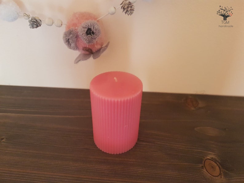 Puede incluir: Una vela rosa en forma de pilar con detalles acanalados verticales. La vela es cil&iacute;ndrica y se encuentra sobre una superficie de madera oscura. Una guirnalda decorativa con cuentas blancas y un adorno rosa y gris cuelga en el fondo.