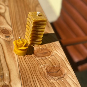 May include: Two yellow beeswax candles, one shaped like a rose and the other a stacked rectangle, on a wooden surface.