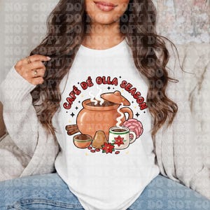 May include: White t-shirt featuring a graphic of a clay pot with steam, a mug, and the text "Café de Olla Season." The design includes cinnamon sticks, a small bowl, and a flower. The overall theme is related to coffee and the fall season.
