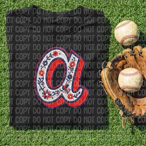 Op de afbeelding: Zwart t-shirt met een rood, wit en blauw bloemenontwerp van de letter "a". Twee honkballen en een honkbalhandschoen op groen gras. Honkbal kleding.