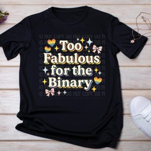 May include: Black t-shirt with the text "Too Fabulous for the Binary" in a retro font. The design includes rainbow hearts, stars, and bows. The shirt is displayed on a light wood surface.