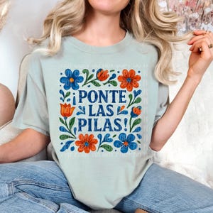 May include: A light blue t-shirt with a floral design and the text "¡PONTE LAS PILAS!" in Spanish.