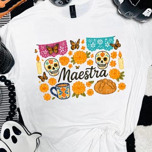 Puede incluir: Camiseta blanca con un diseño colorido del Día de Muertos. El gráfico incluye calaveras de azúcar, flores naranjas, mariposas, velas y la palabra "Maestra". También se representa una taza y pan.
