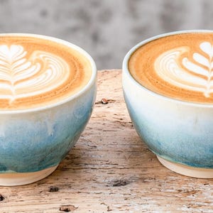 May include: Two ceramic coffee mugs with a light blue and white gradient glaze, each containing a latte with intricate heart-shaped foam art. The mugs feature a rounded handle and a wide rim, showcasing a smooth, glossy finish on the exterior surface.