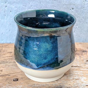 May include: A rounded ceramic mug featuring a glossy glaze that transitions from a deep, dark blue at the rim to a vibrant turquoise in the centre, ending in a smooth, unglazed cream-coloured base with a wide, curved opening and a sturdy foot.