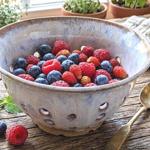 May include: A ceramic colander bowl filled with fresh blueberries, raspberries, and strawberries. The bowl has a speckled, light blue and tan glaze with drainage holes. A gold spoon rests beside the bowl on a rustic wooden surface.