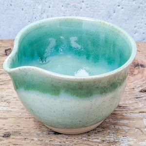 May include: A ceramic pouring bowl with a speckled finish, featuring a light mint green base that transitions into a vibrant emerald green glaze near the rim. The bowl has a distinct spout for pouring and a smooth, rounded interior surface.