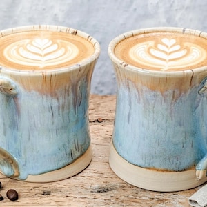 May include: Two ceramic mugs with a flared rim and a handle, featuring a reactive glaze in shades of light blue, cream, and tan. Each mug is filled with a latte topped with a white foam heart-shaped leaf pattern, arranged on a wooden surface.