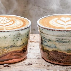 May include: Two ceramic mugs with a reactive glaze finish in shades of cream, sage green, and earthy brown. Each mug contains a hot latte topped with intricate white foam latte art, one featuring a fern pattern and the other a heart design.