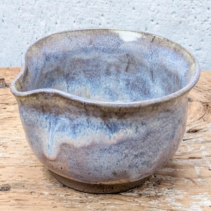 May include: A ceramic mixing bowl with a pouring spout, featuring a reactive glaze in shades of blue, lavender, and cream. The bowl has a rounded body and a dark, unglazed base, showcasing a smooth, glossy finish on the interior and exterior surfaces.
