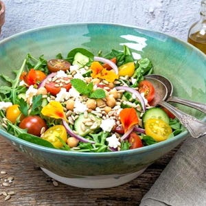 Op de afbeelding: Een kleurrijke salade in een turquoise keramische kom, gevuld met verse ingrediënten. De salade bevat tomaten, komkommers, kikkererwten, rode ui en eetbare bloemen. Zilveren lepels liggen ernaast, met een fles olie en een pepermolen op de achtergrond.