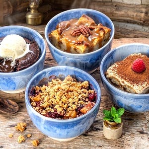 Peut inclure: Quatre desserts individuels dans des bols en céramique bleus sur une surface en bois. Les desserts comprennent un gâteau au chocolat avec de la glace, du pouding au pain avec des noix de pécan, un crumble et un tiramisu.