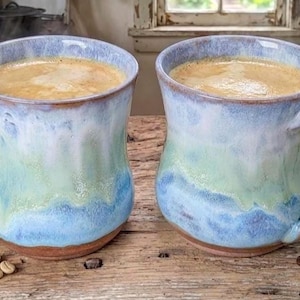 Può includere: Due tazze in ceramica piene di caffè, con una glassa blu e verde. Le tazze hanno grandi manici e sono su una superficie di legno con chicchi di caffè sparsi. Le tazze sono alte circa 10 cm.