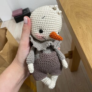 May include: A crocheted snowman toy with a white body, gray overalls, and an orange nose. The snowman is holding a small, white crocheted flower.