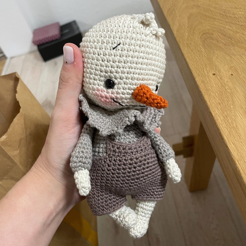 May include: A crocheted snowman toy with a white body, gray overalls, and an orange nose. The snowman is holding a small, white crocheted flower.