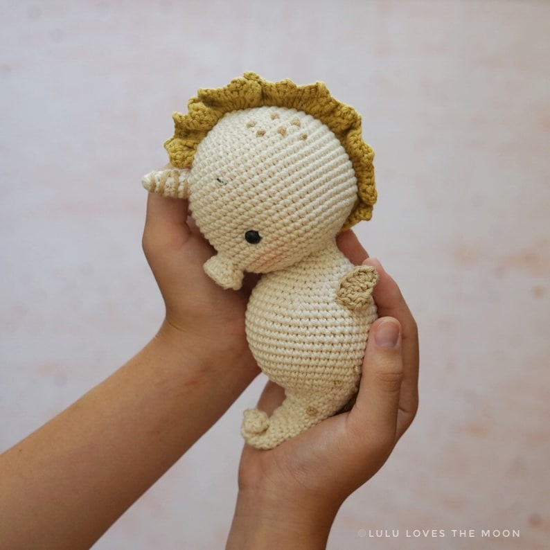 Puede incluir: Un juguete de caballito de mar de crochet con un esquema de color amarillo y blanco. El caballito de mar tiene una aleta amarilla y un cuerpo blanco con ojos negros. El caballito de mar est&aacute; siendo sostenido por las manos de una persona.