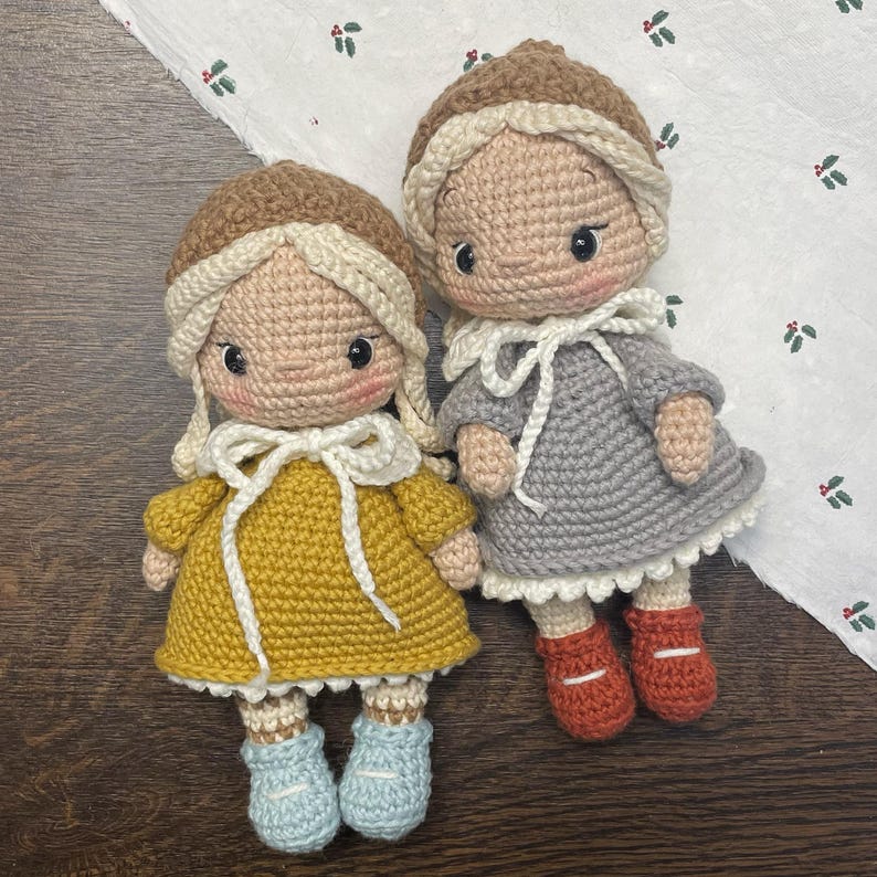 May include: Two handmade crochet dolls. One doll wears a mustard yellow dress and light blue boots, the other wears a gray dress and orange boots. Both dolls have brown hats and blonde hair. The dolls are on a white surface with green and red floral accents.