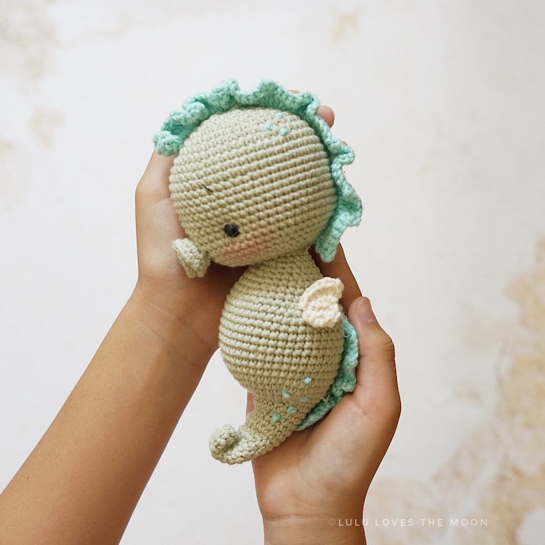 Puede incluir: Un juguete de caballito de mar de crochet con una crin verde claro y blanca, una cola verde claro y una aleta blanca. El caballito de mar est&aacute; siendo sostenido por las manos de una persona.