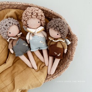 May include: Three crocheted dolls with brown hair and different colored outfits are sitting in a wicker basket. The dolls are wearing overalls and have embroidered details. The basket is lined with a brown fabric.