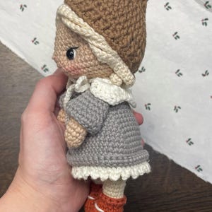 May include: A handmade crochet doll wearing a brown hat with a cream-colored trim, a gray dress with a white lace hem, and orange shoes. The doll has a light skin tone and is holding a small, beige-colored object.