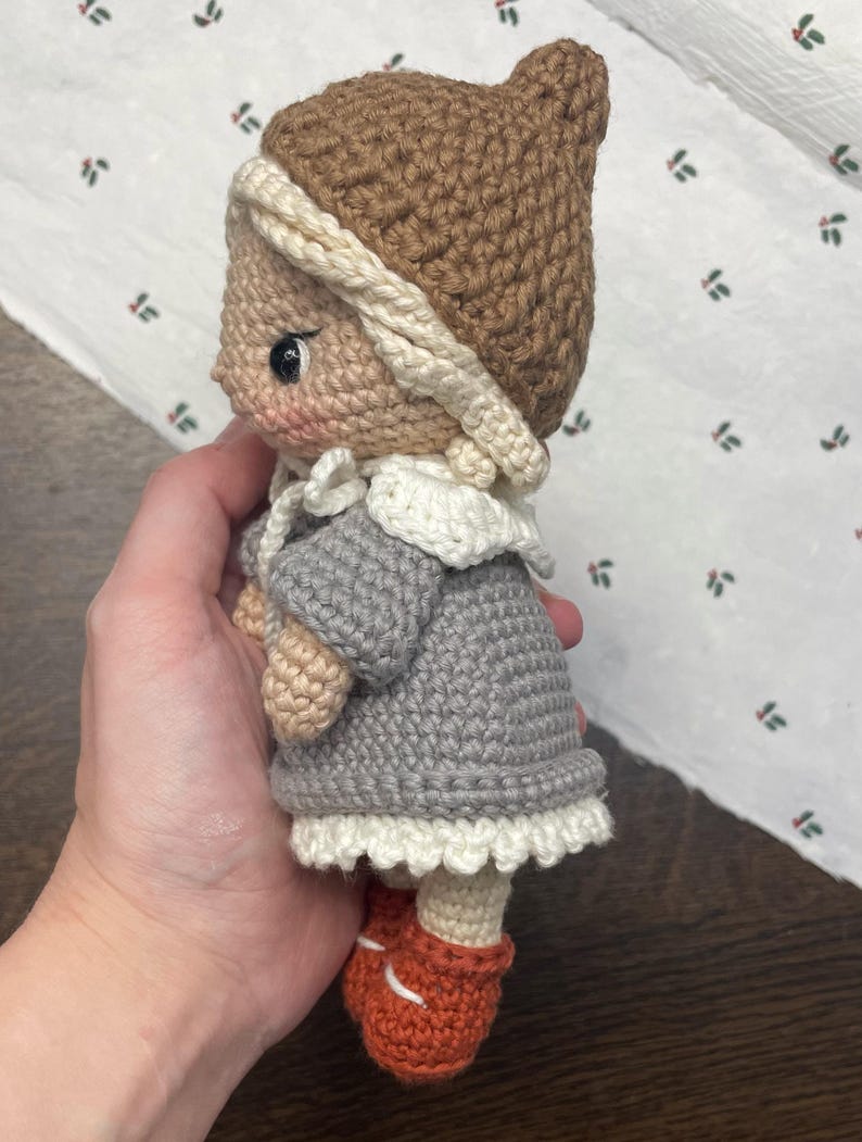 May include: A handmade crochet doll wearing a brown hat with a cream-colored trim, a gray dress with a white lace hem, and orange shoes. The doll has a light skin tone and is holding a small, beige-colored object.
