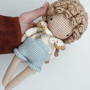 May include: A crocheted doll with light brown hair, wearing a white and yellow crocheted shirt, blue overalls, and a button. The doll has a light brown complexion and is holding its hands together.