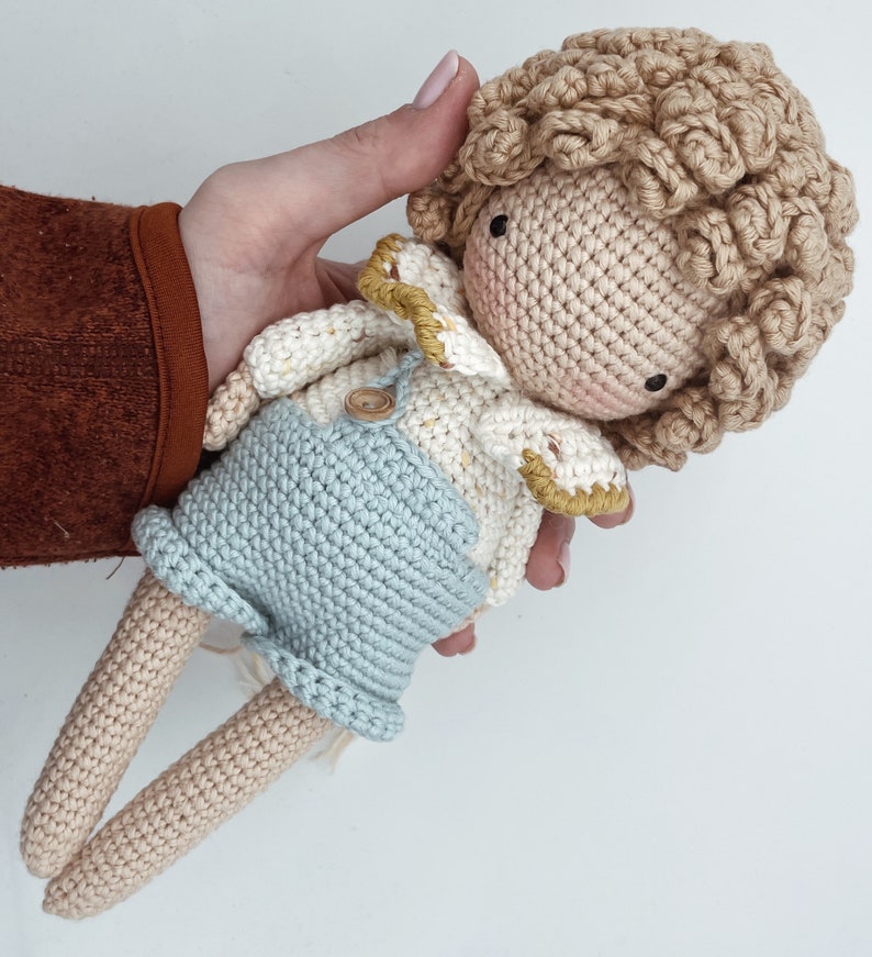 May include: A crocheted doll with light brown hair, wearing a white and yellow crocheted shirt, blue overalls, and a button. The doll has a light brown complexion and is holding its hands together.