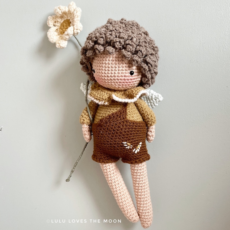 May include: A crocheted doll with brown hair, wearing a brown and tan outfit with a white collar and wings. The doll is holding a small white flower.