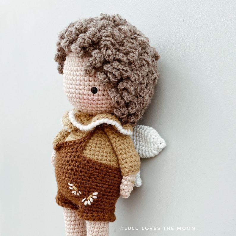 May include: A crocheted doll with brown curly hair, wearing a brown and tan outfit with white accents. The doll has a white crocheted wing on its back.