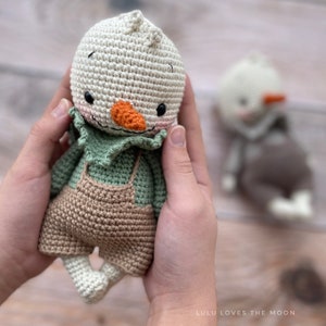 May include: A crocheted snowman doll wearing a green shirt and brown overalls. The doll has a carrot nose, black eyes, and a pink mouth.
