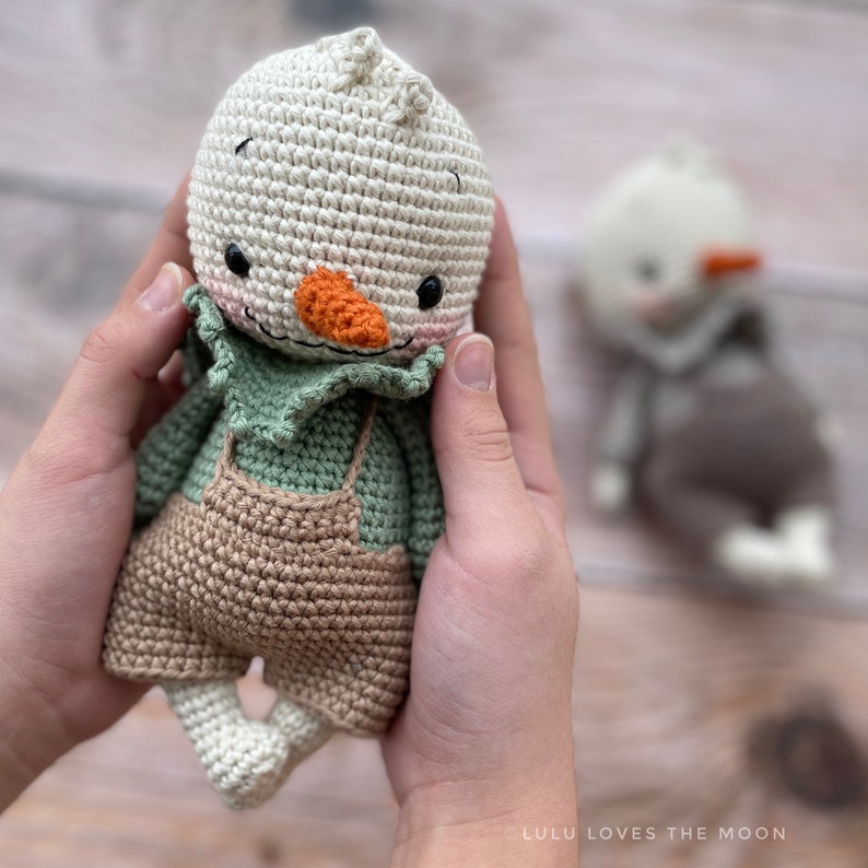 May include: A crocheted snowman doll wearing a green shirt and brown overalls. The doll has a carrot nose, black eyes, and a pink mouth.