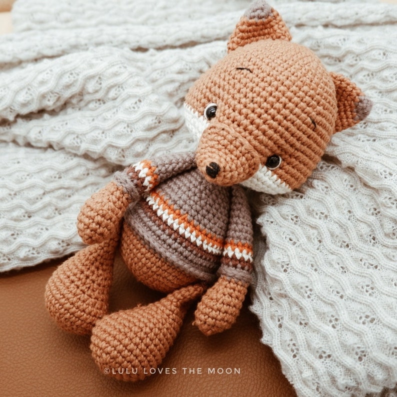 May include: A crocheted fox toy with brown and gray fur, wearing a striped sweater with orange, white, and brown stripes. The fox is lying on a white knitted blanket.