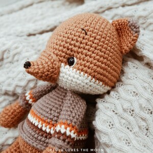 May include: A crocheted fox wearing a brown and orange striped sweater. The fox is lying on a white knitted blanket. The text "LULU LOVES THE MOON" is visible in the bottom right corner of the image.