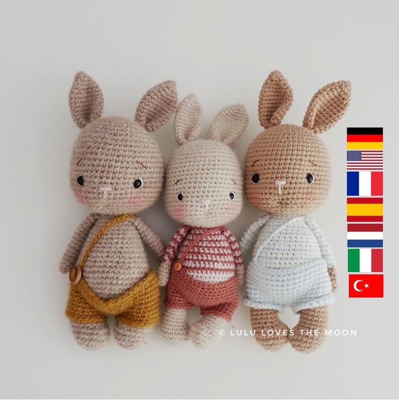 May include: Three crocheted bunny plushies with different colored outfits. The bunny on the left is wearing yellow overalls, the middle bunny is wearing a red and white striped shirt, and the bunny on the right is wearing a light blue shirt.  The bunnies are all beige and have black eyes and noses.