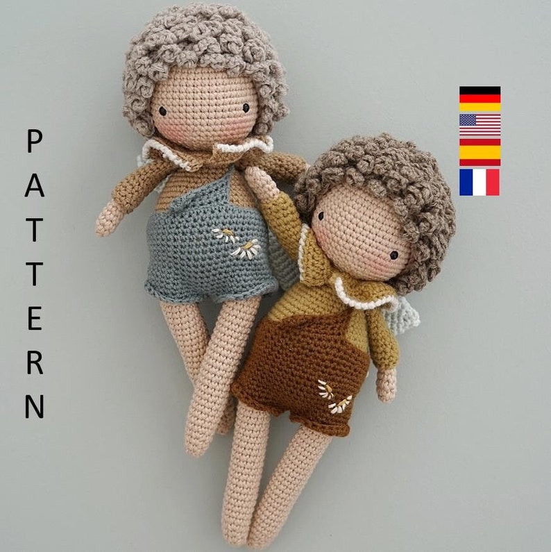May include: Two crocheted dolls with brown and beige clothing. The doll on the left has blue overalls and the doll on the right has brown overalls. Both dolls have curly hair and are smiling.