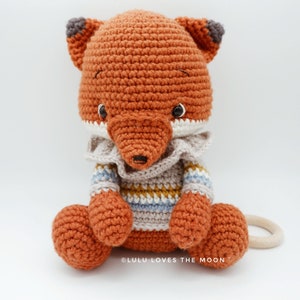 May include: A crocheted fox toy wearing a striped sweater. The fox is sitting with its tail curled around its legs. The toy is made with orange, brown, white, blue, and yellow yarn. The toy is labeled "@LULU LOVES THE MOON".