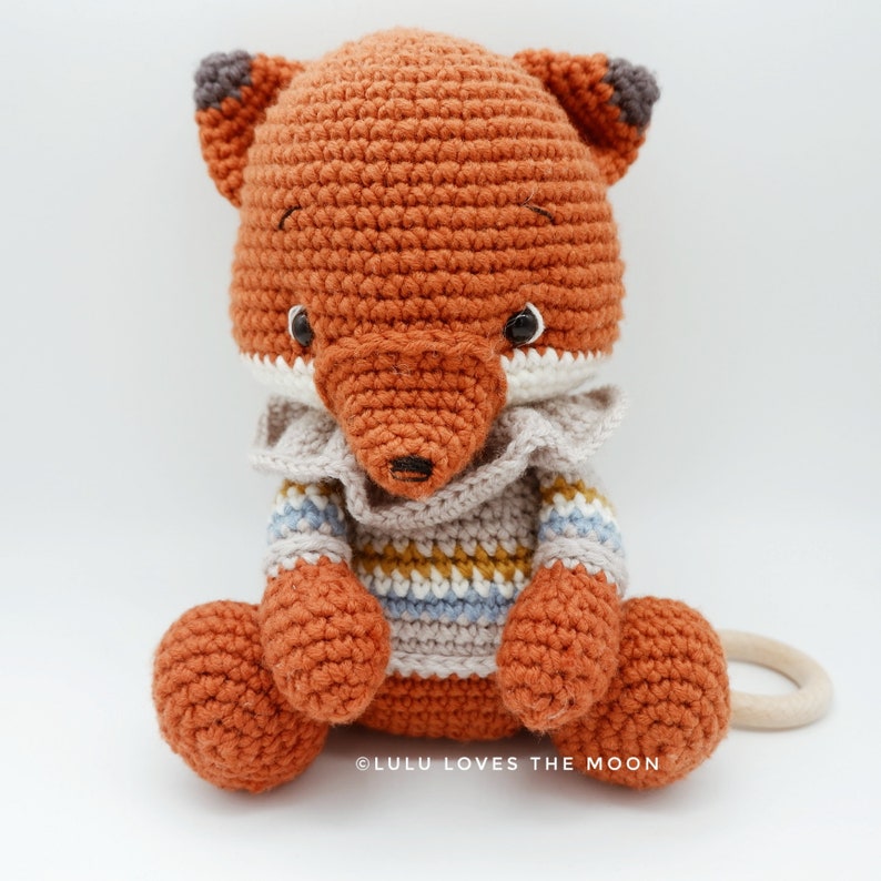 May include: A crocheted fox toy wearing a striped sweater. The fox is sitting with its tail curled around its legs. The toy is made with orange, brown, white, blue, and yellow yarn. The toy is labeled "@LULU LOVES THE MOON".