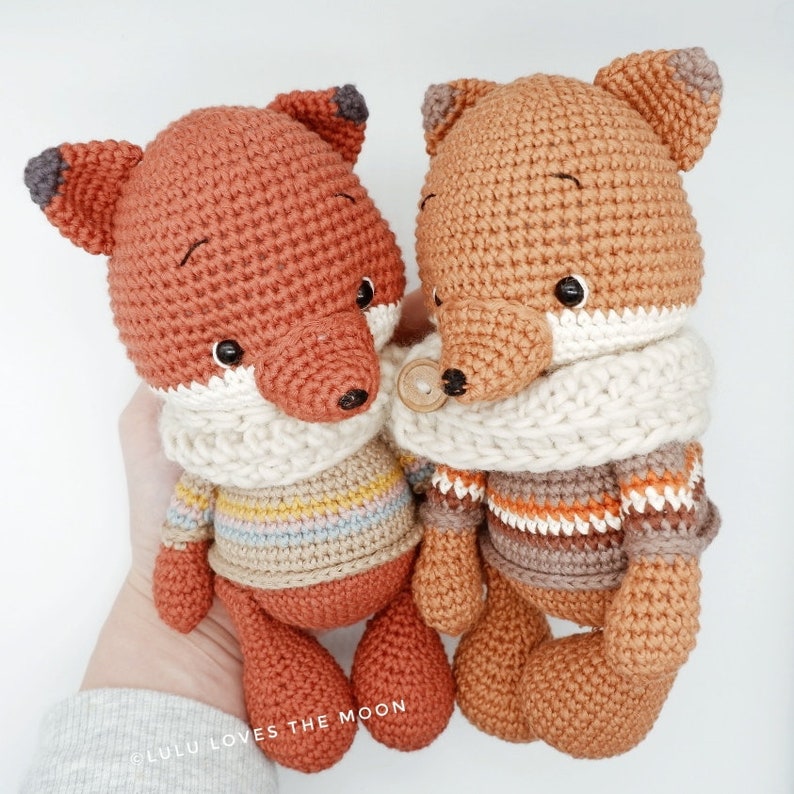 May include: Two crocheted fox plushies wearing striped sweaters and scarves. The fox on the left has a light blue, yellow, and pink striped sweater and a white scarf. The fox on the right has a brown, orange, and white striped sweater and a white scarf.
