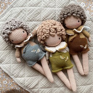May include: Three crocheted dolls with curly hair and embroidered details. The dolls are wearing overalls and have different colored shirts. The dolls are lying on a light brown quilted fabric.