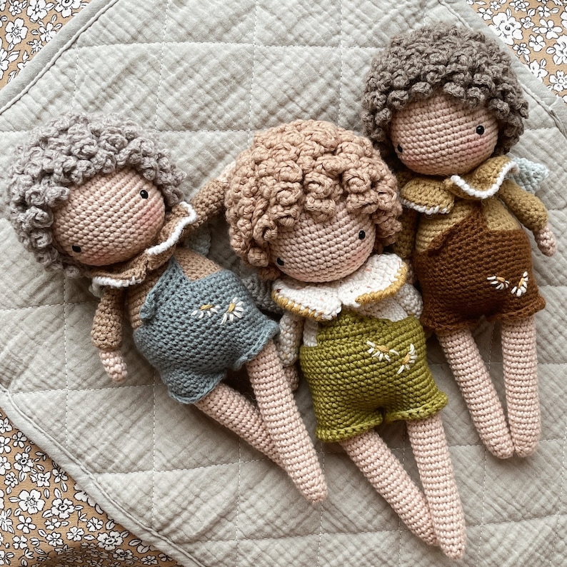 May include: Three crocheted dolls with curly hair and embroidered details. The dolls are wearing overalls and have different colored shirts. The dolls are lying on a light brown quilted fabric.