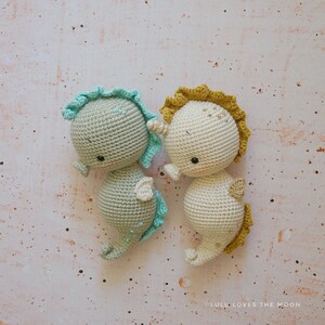 Puede incluir: Dos juguetes de caballito de mar de crochet, uno en verde claro con una aleta turquesa y el otro en beige con una aleta amarilla. Los caballitos de mar tienen ojos negros y miran hacia el lado derecho de la imagen.